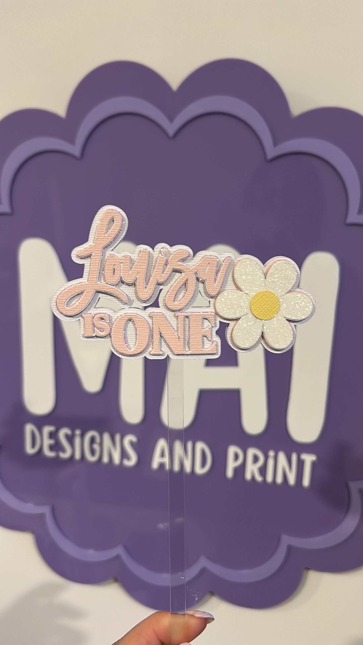 Decorative cake topper with 'Luna is ONE' and a flower design on a purple scalloped border.