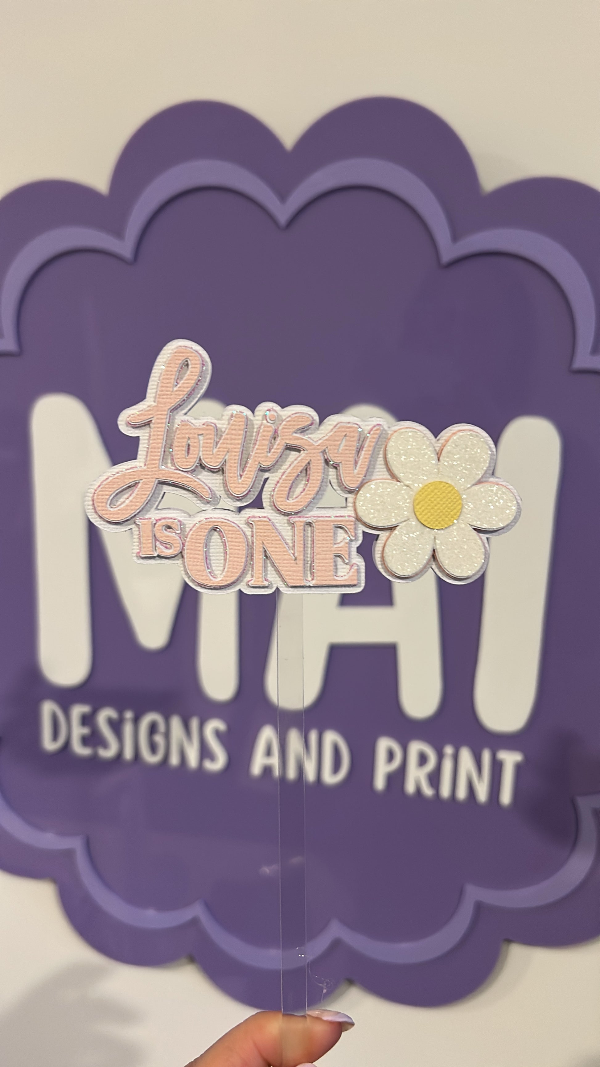 Decorative cake topper with 'Luna is ONE' and a flower design on a purple scalloped border.