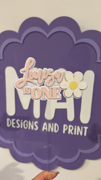 Decorative cake topper with 'Luna is ONE' and a flower design on a purple scalloped border.